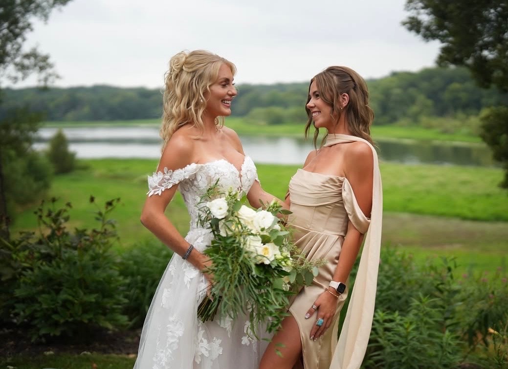 Bride and bridesmaid smiling together outdoors near a lake